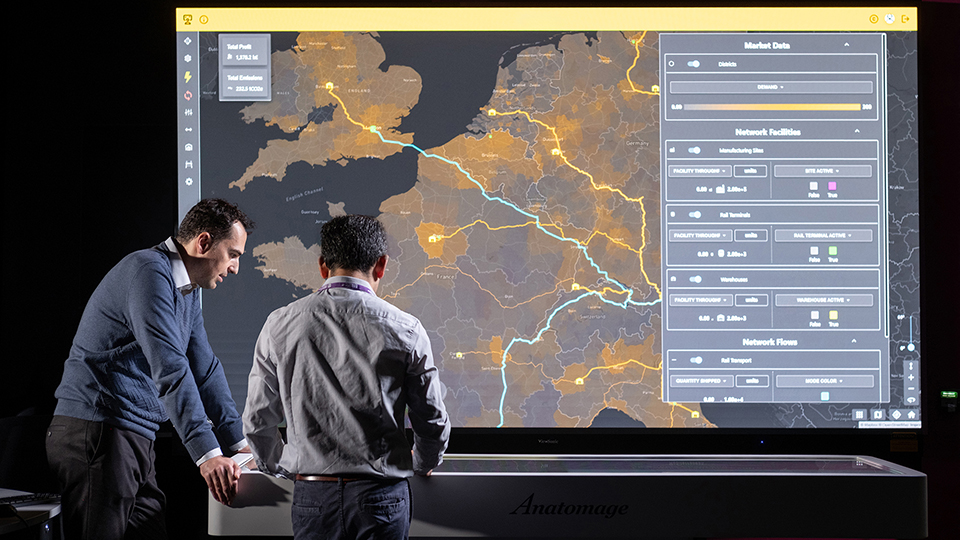 Two people standing in front of a large interactive digital display showing a map-based supply chain or logistics network dashboard, with transport routes, data visualisations and operational controls visible on screen.