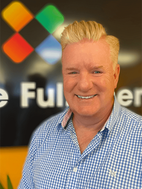 Portrait of Neil Ashworth smiling, wearing a blue checkered shirt, standing in front of a blurred We Are Fulfilment logo