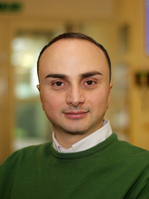 Person wearing a green sweater and white collared shirt, standing indoors in a softly lit environment.