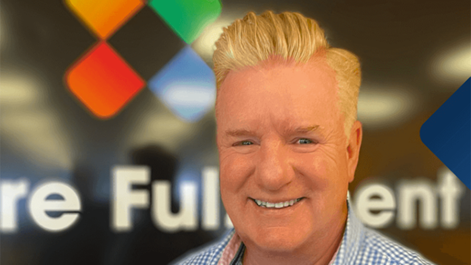 Portrait of Neil Ashworth smiling, wearing a blue checkered shirt, standing in front of a blurred We Are Fulfilment logo