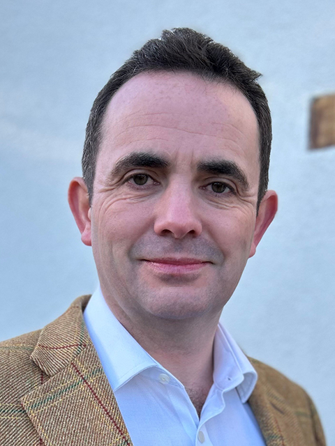 Person wearing a tan plaid jacket and white dress shirt, photographed outdoors against a light-colored background.