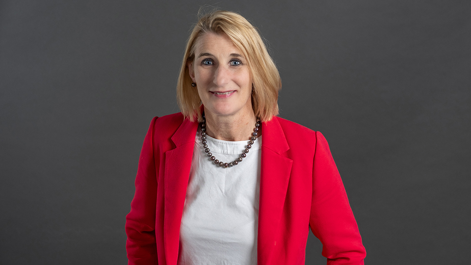 Portrait of Professor Jan Godsell standing against a dark grey background, wearing a bright pink blazer over a white top and a beaded necklace, smiling towards the camera.