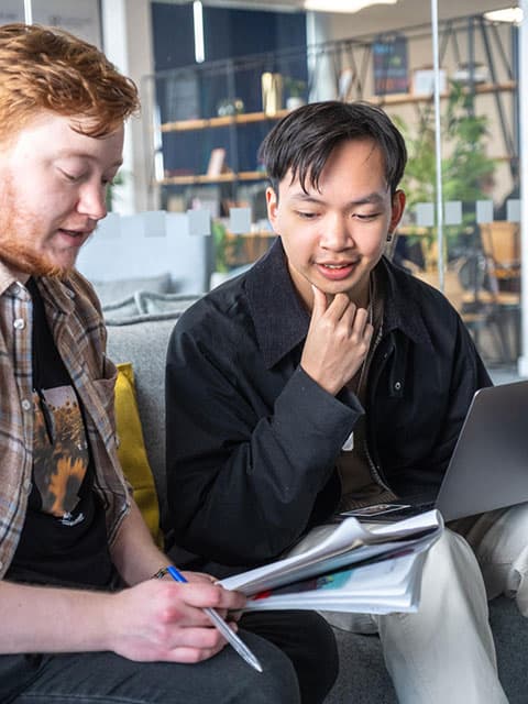 Two students sat down discussing and looking at papers.
