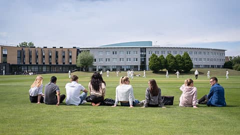 Students sitting on grass watching a cricket match.