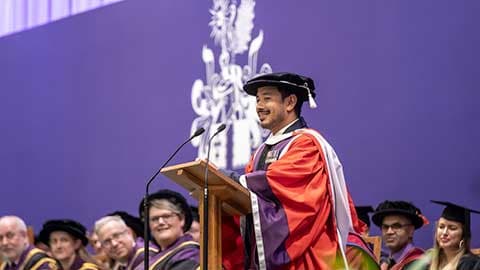 Nims Purja MBE on stage at Loughborough University graduation delivering a speech at a podium.