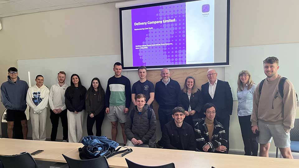 A group of students, lecturers and visiting speakers standing in a row with a presentation screen behind them.