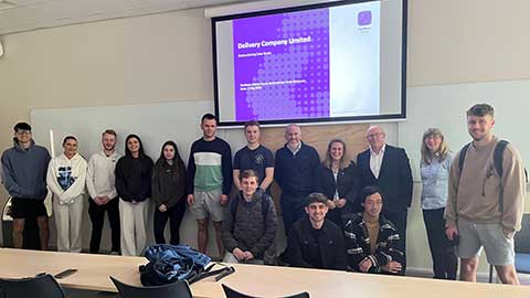 A group of students, lecturers and visiting speakers standing in a row with a presentation screen behind them.