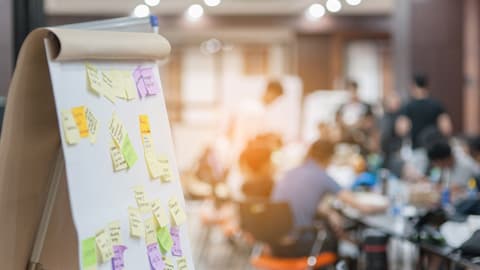 A flipchart board covered in post-it notes, in the background a group of people are sat around a table.