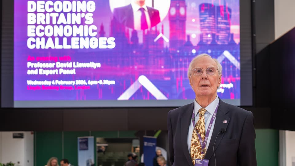 Professor David Llewellyn stands in front of a screen promoting his lecture on Britain's economy.