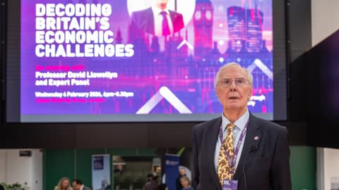 Professor David Llewellyn stands in front of a screen promoting his lecture on Britain's economy.