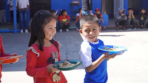Two school children in Guatemala carrying plates of food in the playground.