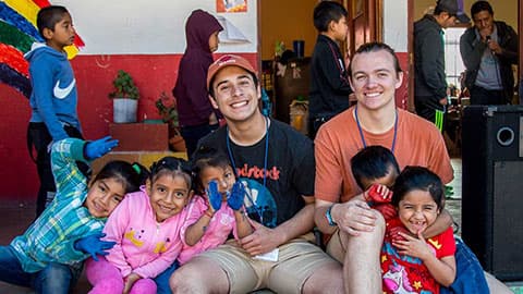 Two adult volunteers with five school children smiling and sitting on a step outdoors. The children's hands are coloured with paint.