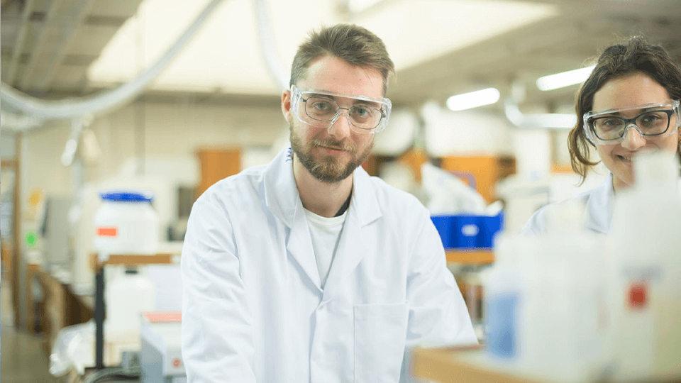 male student in lab coat and goggles looking at camera with female student in the background