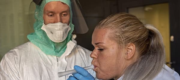 a woman breathes into a testing tube, a man wearing PPE watches