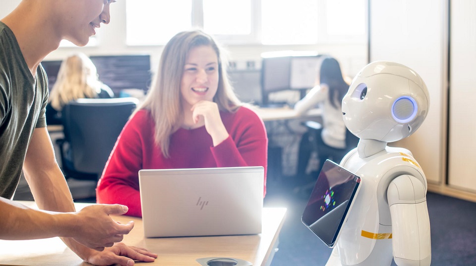 Female student Emma sat down at a table smiling at humaoid robot Pepper. Male student can be seen part in the image leaning on the table.