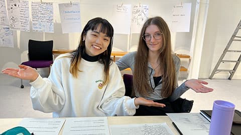 two students holding their hands out at the sides, there are notes covering the desk in front and both are smiling