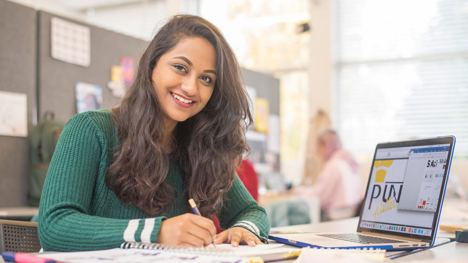 A student smiling at the camera in front of a laptop