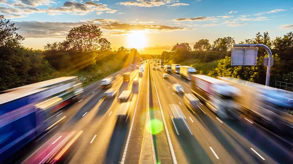 A bustling multi-lane highway filled with fast-moving cars and large trucks driving toward a bright, glowing sunset.
