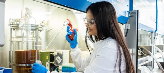 person with long brown hair wearing lab goggles, coat and gloves holding a test tube