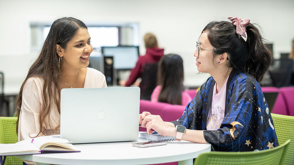 Two female students sitting side by side around a table having a conversation whilst using a laptop featuring other students working in the background. 