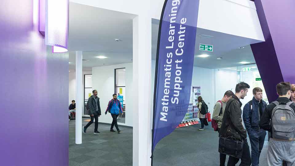 Wide angle showing students walking inside a building with a flag featuring up front and centre reading 'Mathematics Learning Support Centre'.