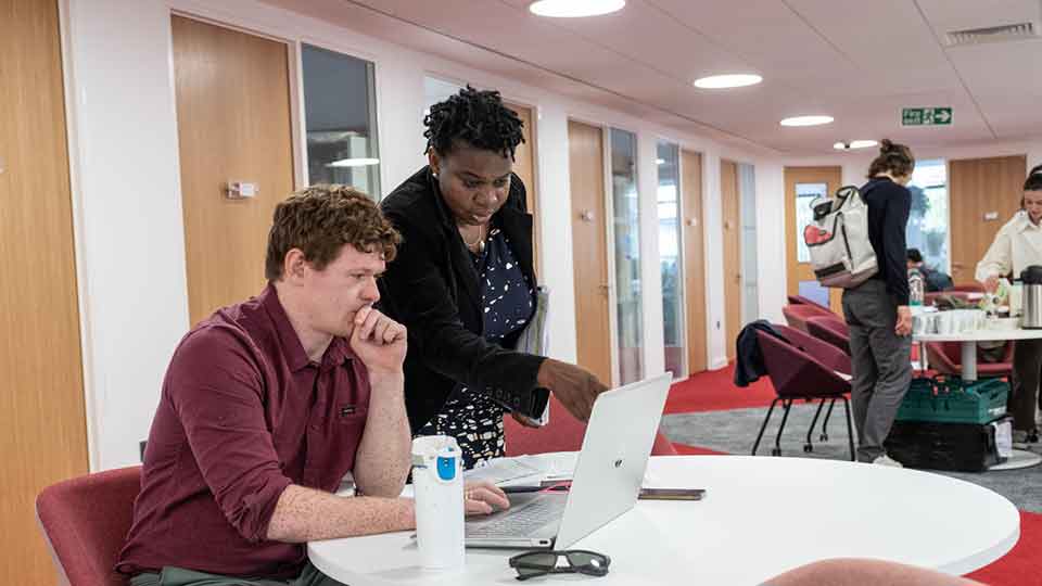 Dr Sheryl Williams leaning of the shoulder of a student showing them something on a laptop screen who is sat at a table looking directly at the laptop.