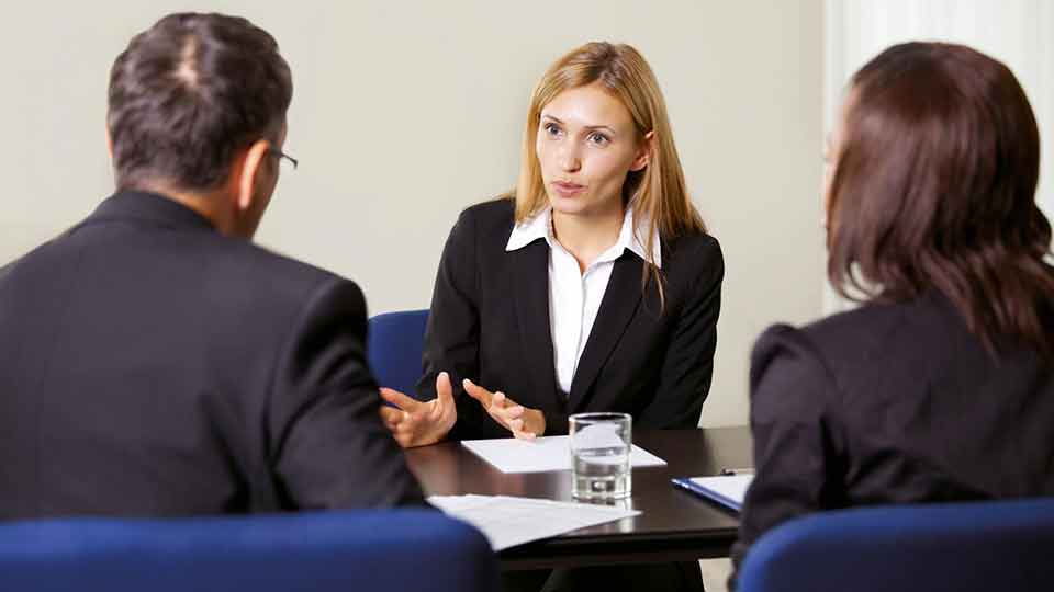 Three people sat around a table having a conversation, one woman is using her hands whilst speaking, whilst the other two participants can be seen from behind, listening to her.