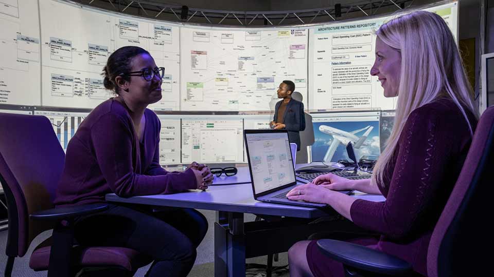 Two female students sitting at a table facing each other smiling, whilst in conversation. A PhD student is featured in the background looking at data on an interactive screen. 