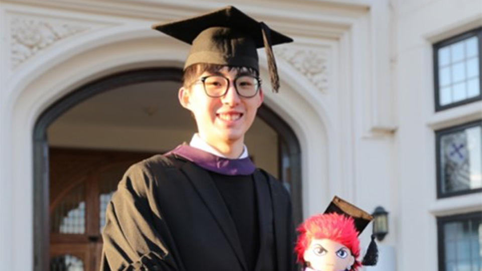 Jonathan stood outside the Hazelrigg building on campus at Loughborough, wearing a graduation gown and hat, smiling, looking directly at the camera.