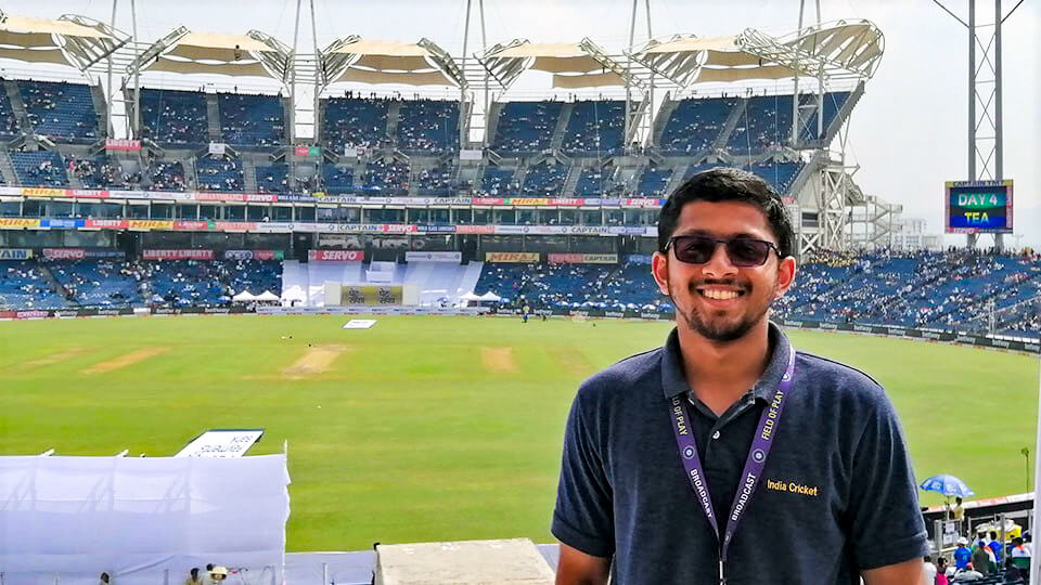 Graduate Ebbin Binny standing smiling in the stalls on a cricket ground showing the cricket pitches and opposite stalls in the background. 