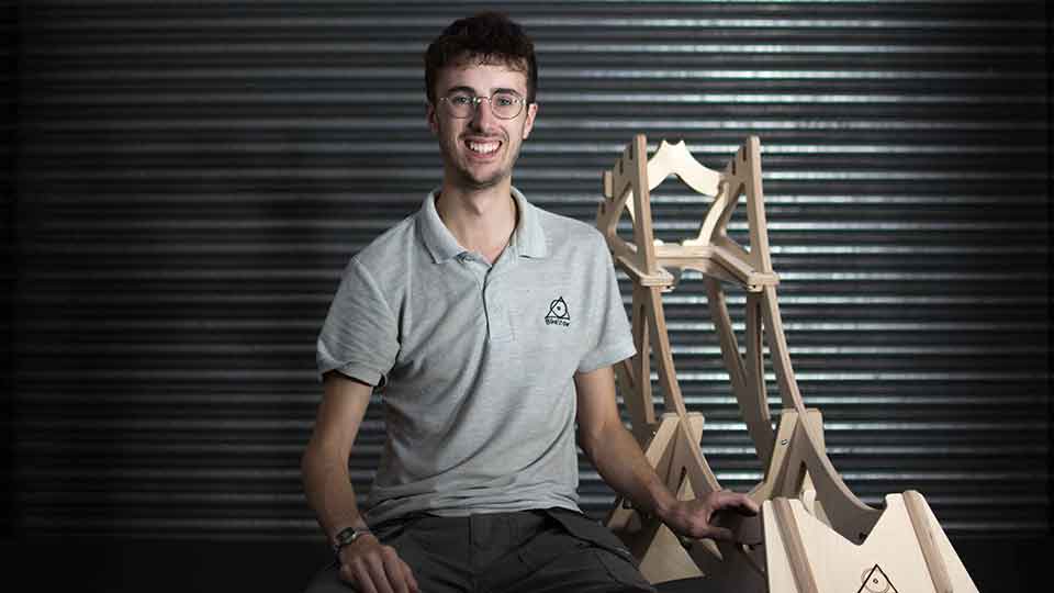 Student George sitting whilst looking and smiling directly at the camera with his right hand on a wooden bike storage unit.