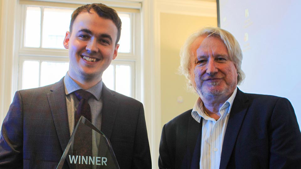 Graduate Guy King standing in a photo holding a glass trophy winner's prize with an academic stood to his right-hand side.