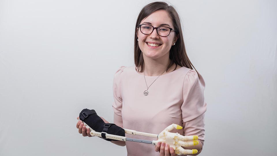 Product Design Engineering graduate Kate Walker headshot, smiling and looking directly into the camera whilst holding a prosthetic arm.