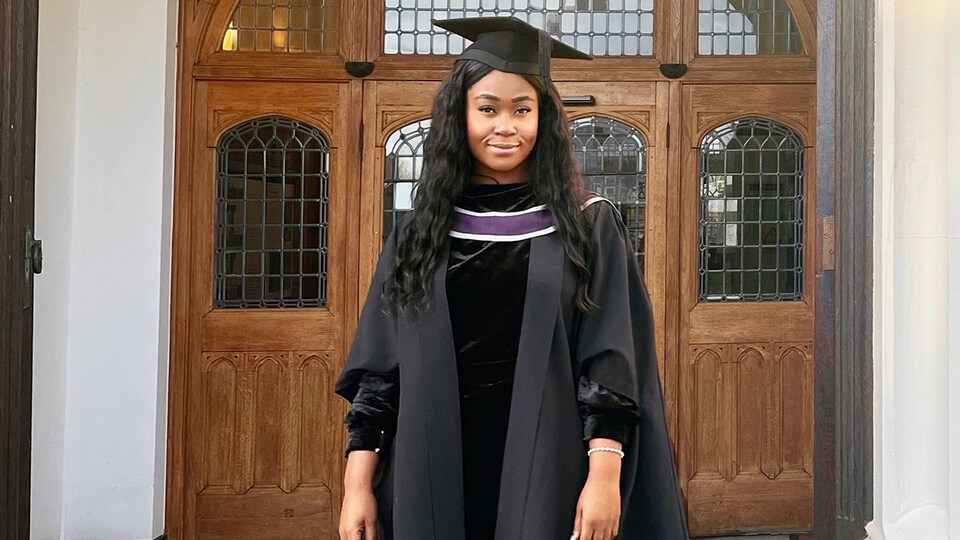 Leah standing in front of the entrance of a building on campus at Loughborough whilst wearing her graduation gown and hat, smiling, and looking directly at the camera. 