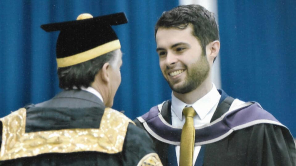 Michael wearing at his graduation wearing a robe whilst smiling, looking at a professor who is photographed from behind wearing a graduation hat and robe.