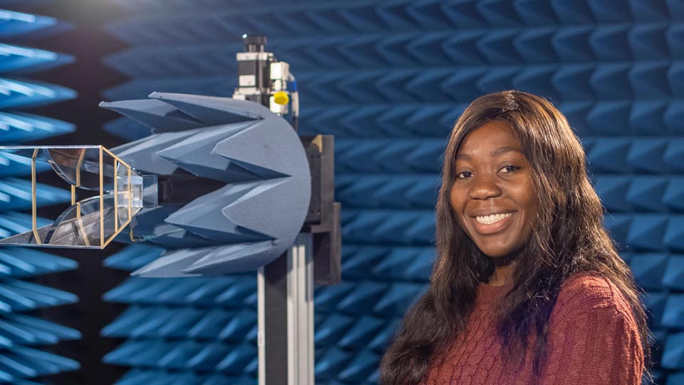 Headshot of Olamide standing in an anechoic chamber next to a piece of equipment.