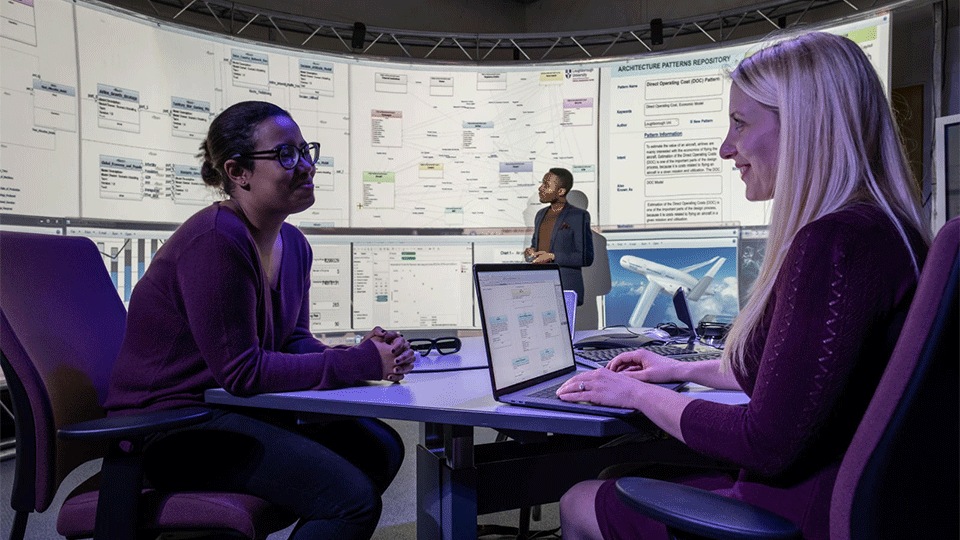 2 women facing each other smiling with big digital screen in background