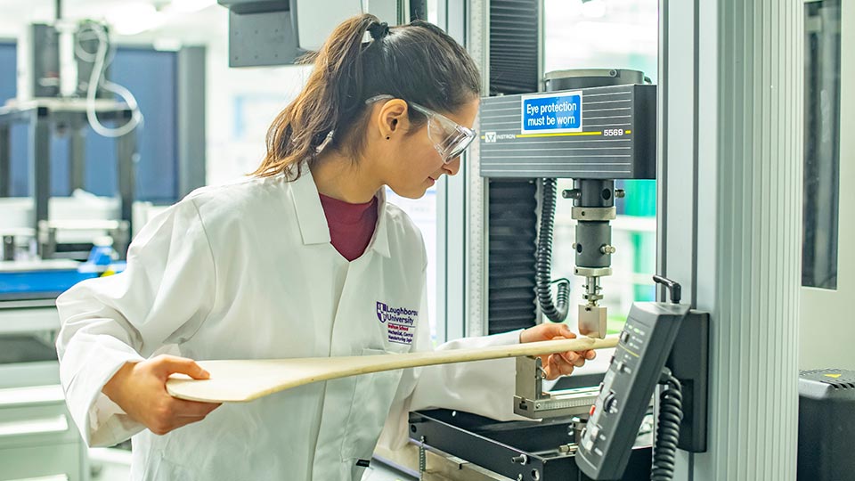 Side shot of Vedanti in the Sports Technology lab holding a piece of wood whilst using a piece of equipment. Vedanti is wearing PPE such as goggles and overalls. 