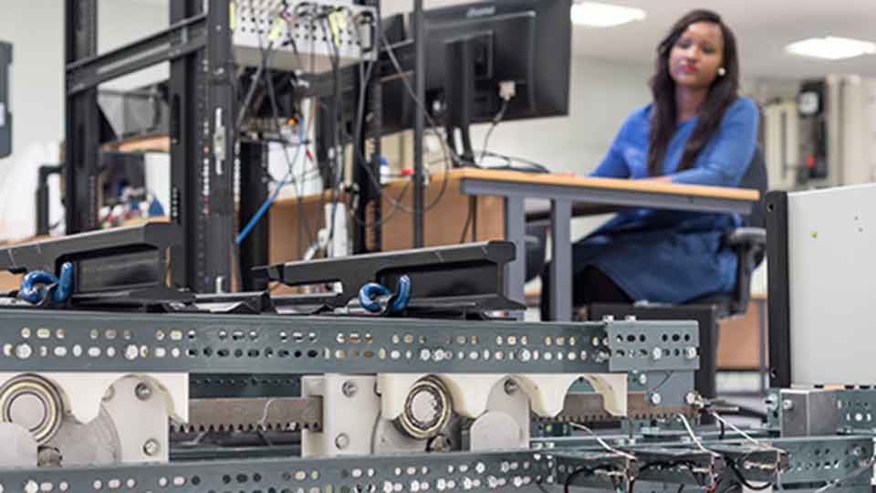 A close-up shot is focused on cables and electronic systems. A female is sitting in the background at a computer, looking down towards the equipment.