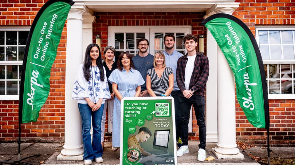 A group of 7 standing outside, in front of a house, all smiling and looking at the camera. Green and white flags are positioned either side of them which read 'Sherpa- one-to-one online tutoring.' 