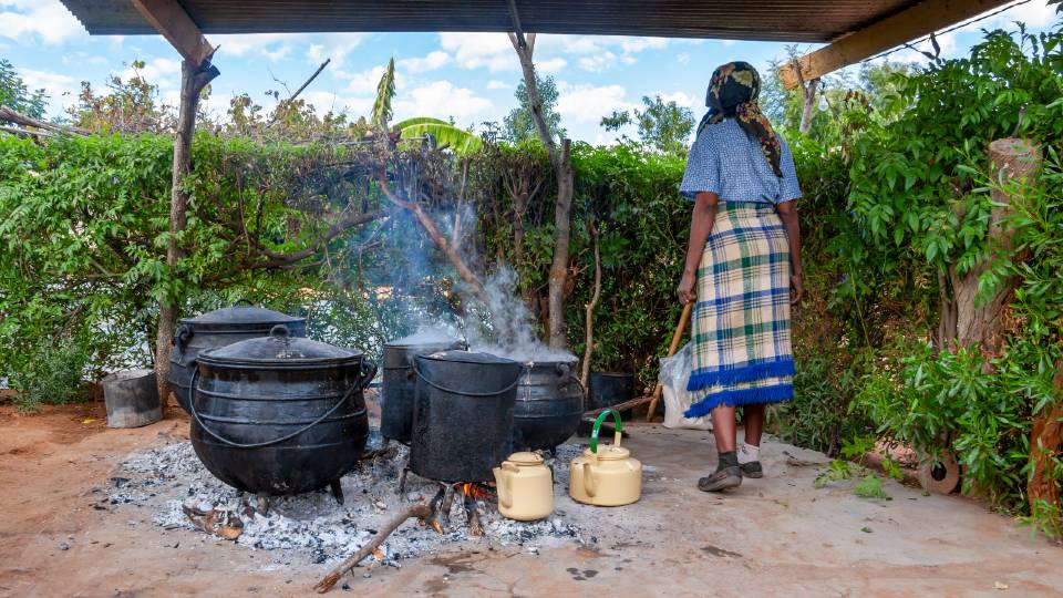 A full body shot of a human standing next to 5 large cast iron cooking pots and two smaller cast iron kettles with their back to the camera. 