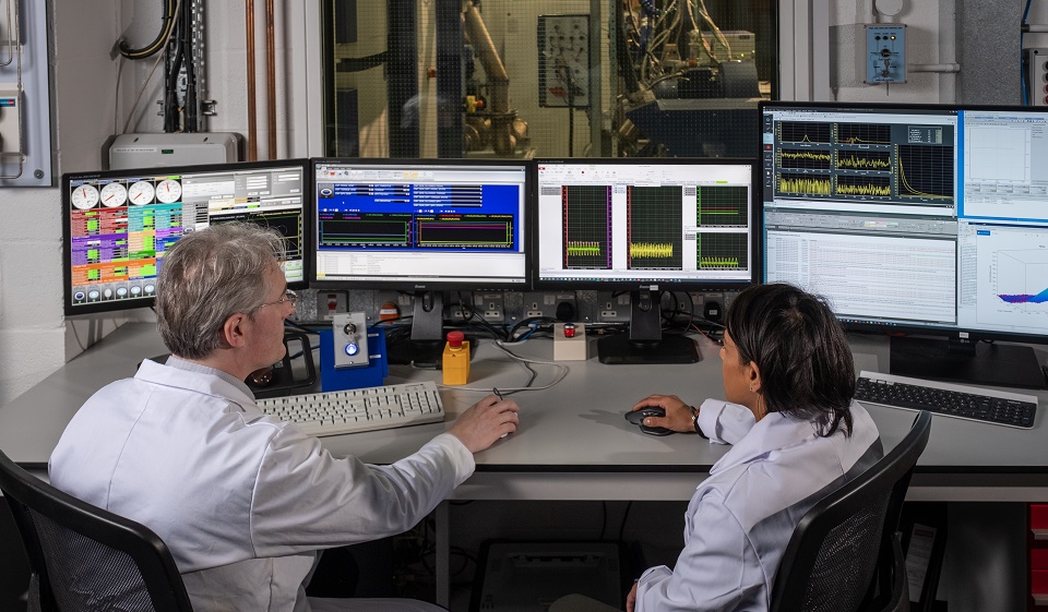 Two academics sat in a lab where monitors are on the worktop.