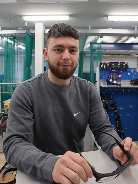 Portrait image of Derry, smiling, looking at the camera whilst holding a pair of sports sunglasses.