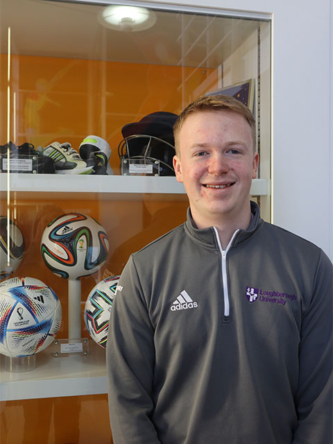 Portrait shot of Ieuan, standing in front of a clear glass cabinet which contains footballs. 