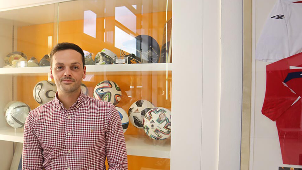 Dr Jon Farmer standing in front of a glass cabinet showing footballs and sports equipment in the Sports Technology Institute whilst wearing a red chequered shirt. 