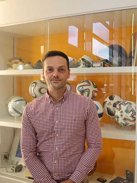 Portrait shot of Jon standing in front of a glass cabinet where footballs are displayed in the Sports Technology Institute