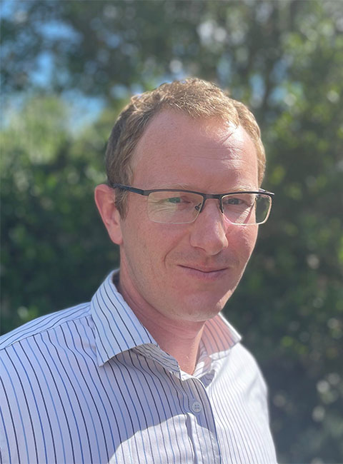 Headshot of Nick, smiling looking at the camera outside where the sun is shining. 