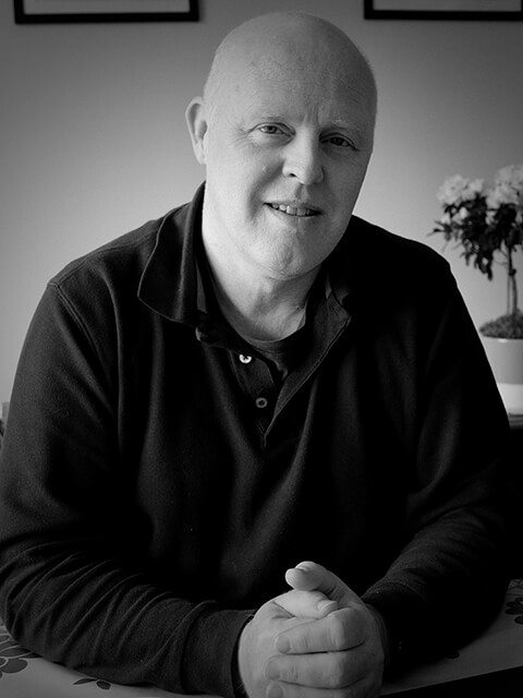 Black and white portrait image of Paul sitting at a table looking directly at the camera, smiling. 
