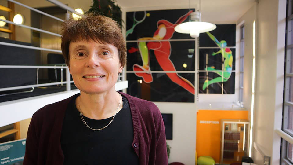 Dr Steph Forrester standing on the stairs in the Sports Technology Institute, smiling with artwork of colourful digital assets of humans playing various sports in the background.
