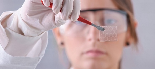 a woman wearing a lab coat and safety glasses holding graphene film with tweezers